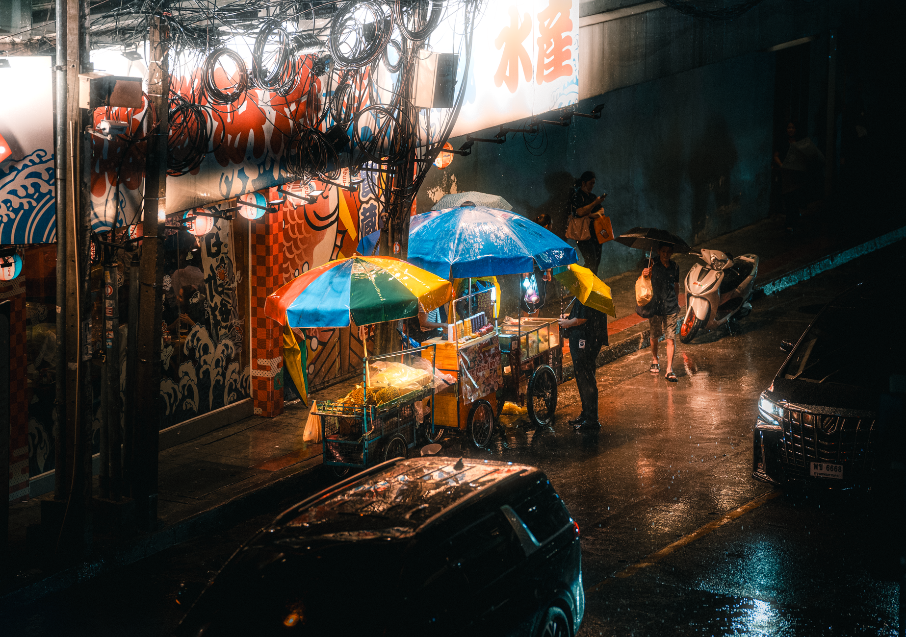 Lively street food stalls under colorful umbrellas in rainy Bangkok at night.